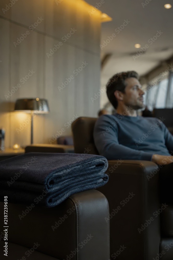 Fototapeta premium A man is comfortably settled in a cozy lounge area, surrounded by a pile of soft blankets. The warm lighting creates a serene atmosphere as he gazes thoughtfully out the window, lost in his own reflec