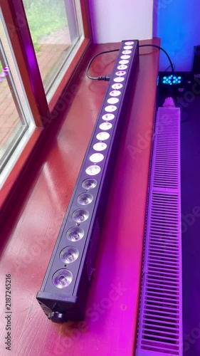 A linear LED light and speaker on a wooden windowsill next to a radiator, set against a window; multicolored LEDs are running and changing color. Vertical background with copy space