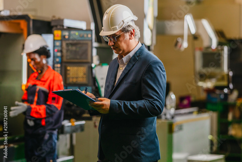 Senior engineer with clipboard inspecting CNC machines and machined part in parts factory