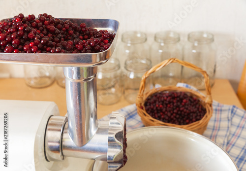 Making lingonberry homemade jam. Grinding ripe red berries in an electric meat grinder. Woven basket of berries and jars on the kitchen table. Preparing winter supplies. Close-up view
