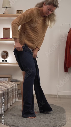 Young woman in a sweater putting on tight jeans in her living room at home, getting dressed and preparing to go out.