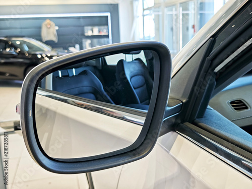 Side mirror of a white Chinese car reflecting rear seats inside a modern automobile dealership showroom, close-up of vehicle exterior detail
