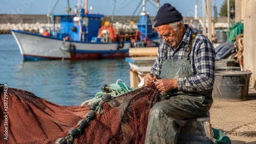 Senior fisherman repairing fishing net by the harbor with boats in the background on a sunny day