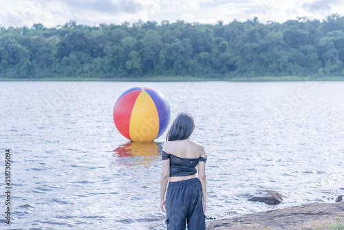 A young Asian woman standing with her back to the camera by a serene reservoir, looking out across the water wearing a stylish black outfit on a bright sunny summer day, with a colorful floating ball.