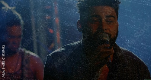 Singing lead singer holding mic and cable on dim stage, with bandmate in sleeveless patterned top
