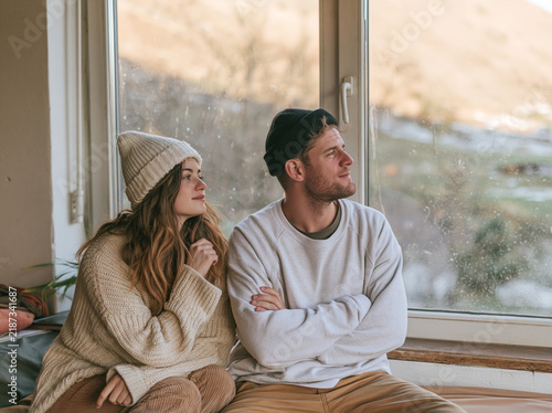 Young couple in warm sweaters and beanies sitting by the window in winter