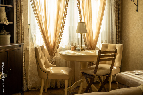 Elegant classic interior with a round dining table, upholstered chairs, decorative cabinet and soft sunlight coming through sheer curtains.