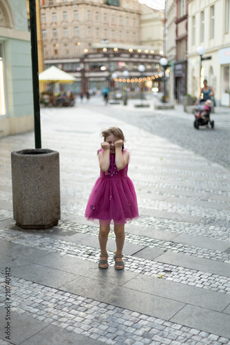Fotografie young girl covers face with both hands while standing on cobblestone pedestrian street, soft blurred city buildings