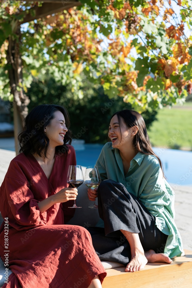 Fototapeta premium Two women laughing with glasses of wine by the pool under autumn vines for warm social background and lifestyle banner design
