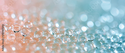 Close-up macro shot of glistening clear gel balls or water beads arrayed across a surface, featuring a dreamy bokeh background gradient transitioning from soft peach to tranquil blue,