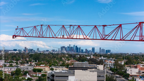 Aerial view of a red construction crane jib extending over the Los Angeles skyline with residential buildings and trees in the foreground in Los Angeles, California, United States.