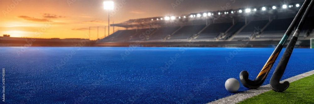 Obraz premium Field hockey sticks and ball on blue artificial turf pitch at sunset. Empty stadium with floodlights in background. World field hockey championship sport game competition. Banner with copy space