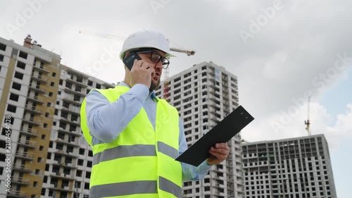 Professional engineer communicating via smartphone while checking papers at construction site. Bearded architect supervising building work