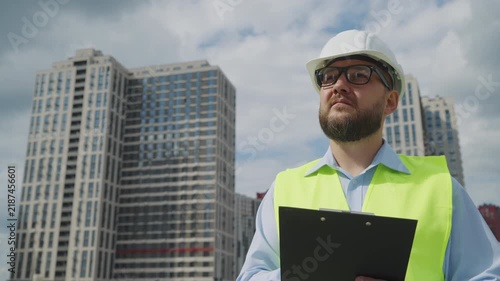 Bearded architect in safety vest holding clipboard and looking at skyscraper. Professional inspector supervising residential building work