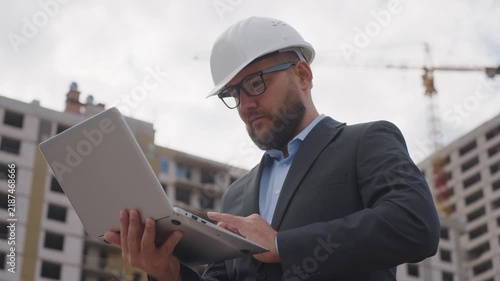 Successful architect in suit and white helmet working on laptop at building site. Professional manager checking project data outdoors
