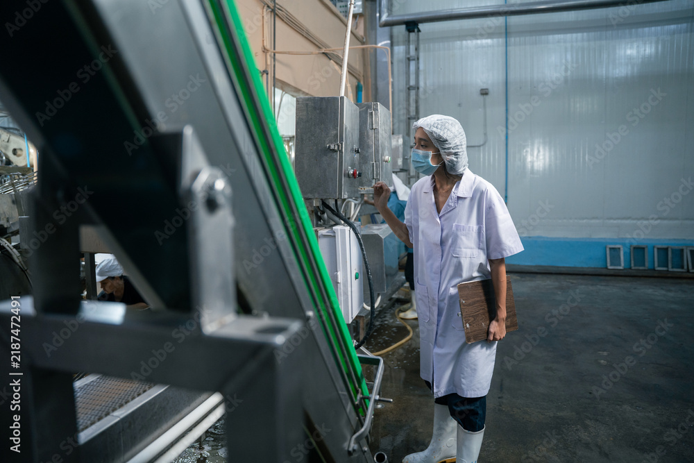 Fototapeta premium A food plant worker wearing a protective hairnet and face mask operates machinery control panels inside a large manufacturing factory with metal equipment and concrete floors.