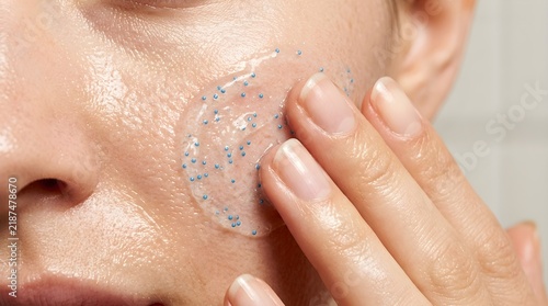 Close up view of woman fingers applying clear gel scrub with tiny blue exfoliating particles on clean cheek skin for deep pore cleansing and exfoliation.