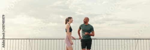 Male and female athletes are talking outdoors on the seafront  walking bridge. Happy fit people are cheerfully talking while working out. Minimalism sport aesthetic. Panoramic