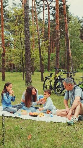 A cheerful family shares a picnic in a pine forest after biking, surrounded by nature, enjoying sandwiches, fruit, and time together.