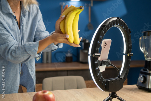 Food blogger livestreaming on social media, talking about healthy diet and showing bananas to camera using smartphone in kitchen. healthy food, diet, social media and blogging.