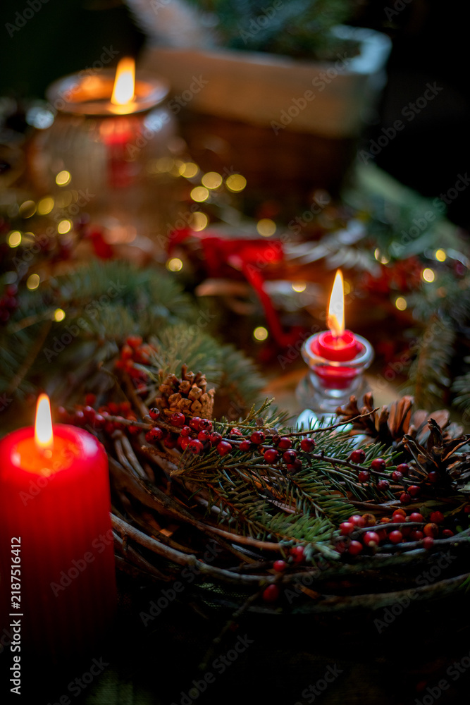 Fototapeta premium Dark and cozy Christmas arrangement with red candles, evergreen branches, red berries and pinecones. Warm candlelight and soft bokeh create a moody, intimate festive atmosphere.