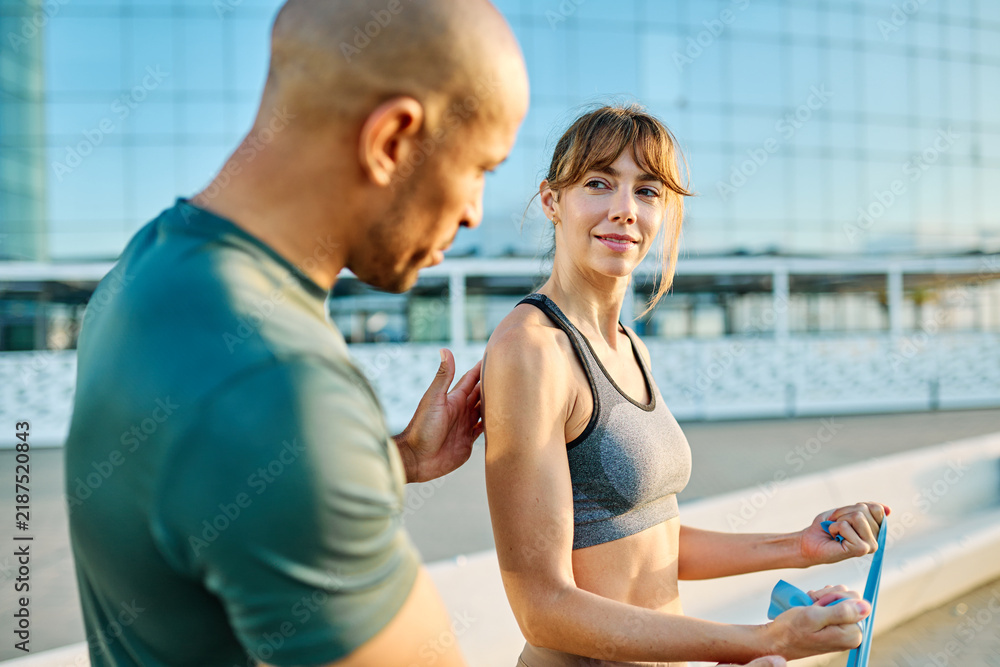 Fototapeta premium Portrait of a happy beautiful woman exercising with resistance band and stretching with trainer coach support outdoors