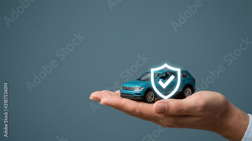 photograph depicting a modern vehicle protection concept in a refined studio setting. A carefully groomed hand enters from the lower right and presents a detailed miniature turquoise-blue SUV 