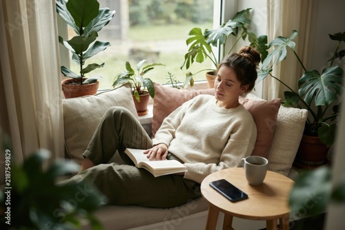 Young woman reading a book while relaxing on a cozy window seat with indoor plants