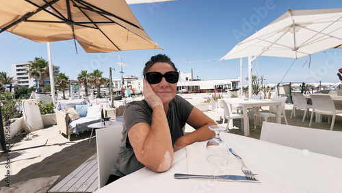 A woman wearing sunglasses sitting at an outdoor restaurant table near a beach.