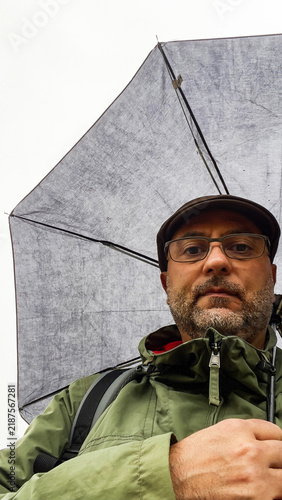 A man wearing glasses, a flat cap, and a green jacket holds an open umbrella outdoors.