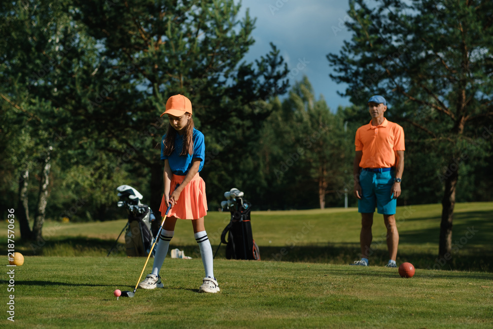 Obraz premium Young girl practices golf swing on a sunny day while an adult watches on a lush green course surrounded by trees in the background