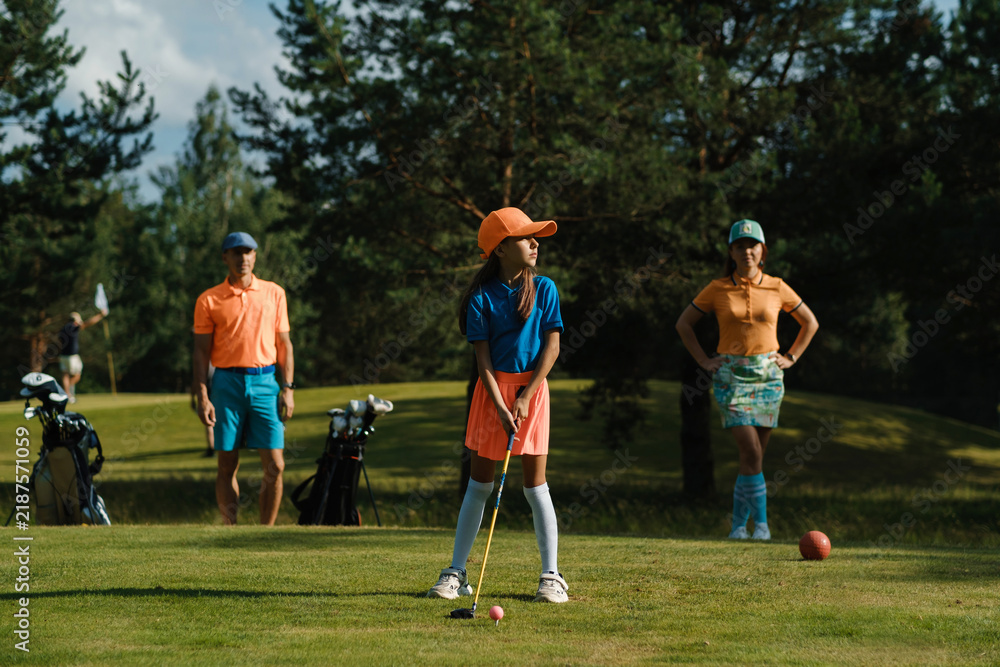Obraz premium A young girl prepares to hit a ball on a sunny golf course surrounded by her parents, playing a family game of golf on a weekend morning