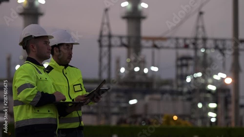 Professional engineers using a digital tablet at a petrochemical refinery or oil and gas power plant during twilight. Concept of energy industry, petroleum infrastructure, and industrial teamwork.