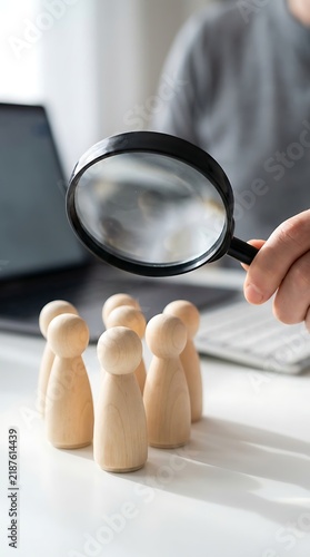 A black magnifying glass held by a human resource recruiter over several wooden figures representing employees on a bright white office table near laptop.