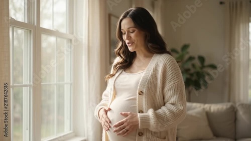 Pregnant woman standing by a sunlit window, gently stroking her belly with love and anticipation.