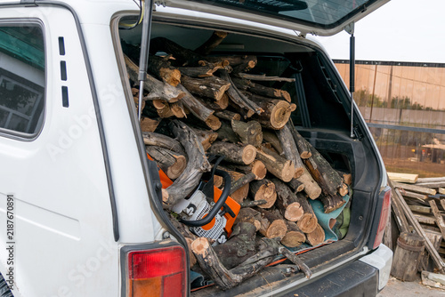 Obraz na płótnie Side view of a white SUV open back door packed with chopped firewood logs and a portable petrol chainsaw