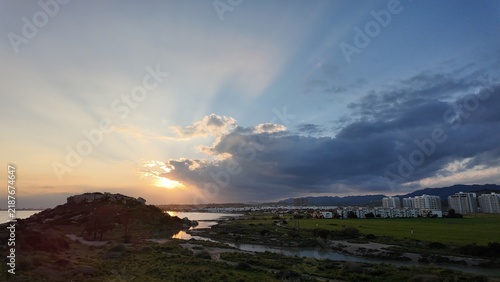 Sunset over hills and the city: rays break through the clouds, reflect in the winding water and create contrast between nature, buildings and mountains on the horizon.