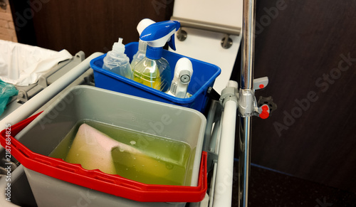 Professional cleaning trolley with plastic buckets, spray bottles, detergents, and cleaning cloth in dirty water. Housekeeping and janitorial service equipment in a hotel or office corridor.
