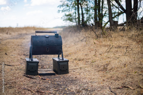 Improvised barrel smoker grill emitting smoke on a dirt trail beside dry, drought-stricken grass and woodland, highlighting open-flame fire risk in tinder-dry conditions.