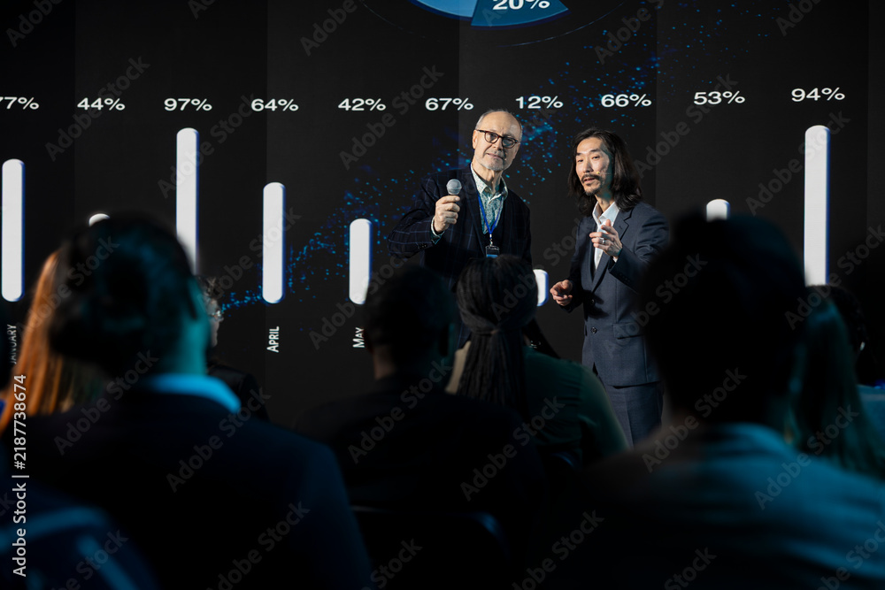 Obraz premium Two presenters of different ages deliver a speech at a business conference while professionals attend an international summit in an auditorium.