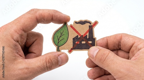 Hands holding a cutout symbol of a leaf next to a factory with a red cross over it