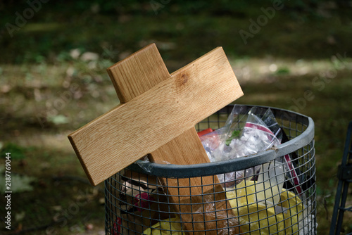 A simple wooden cross disposed of in an outdoor wire mesh garbage can along with plastic waste