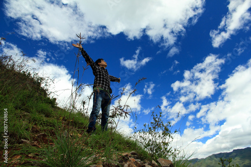 Foto An adult stands on a grassy hillside with arms outstretched, looking up at a bright blue sky with white clouds, surrounded by natural terrain and vegetation