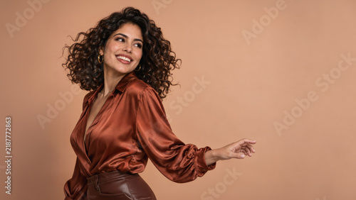 A smiling woman with dark curls poses in a rust satin blouse, brown leather skirt and gold hoop earrings against a warm peach backdrop.
