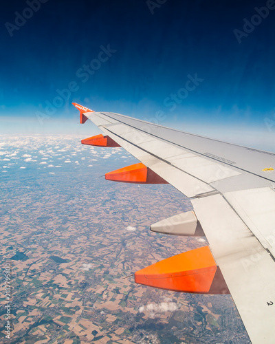 An aerial view from an airplane window captures the wing of an easyJet aircraft soaring above a patchwork of farmlands and scattered clouds, representing the perspective of a passenger during a flight