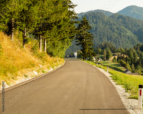 A winding asphalt road leads through a lush, forested mountain landscape towards a small church and village in the distance, capturing the serene beauty of the rural Alpine countryside.