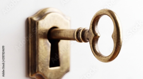 A vintage brass skeleton key inserted into a decorative keyhole on a white wall surface with inserted matching ornate classic antique brass with keyhole plate mounted