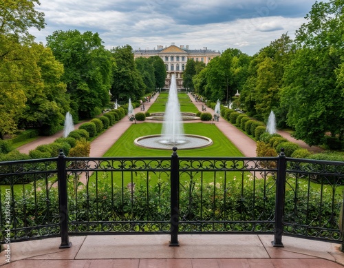 Grand fountain in a landscaped garden with symmetrical pathways, lush greenery, and historical building in the background under a cloudy sky