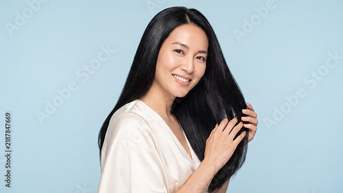 Woman with long black hair smiles gently while gently touching her smooth strands against a plain light blue background in a studio setting.