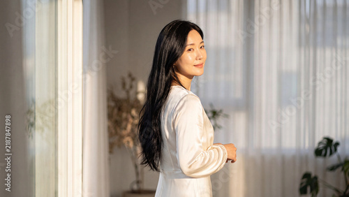 Female model with long dark wavy hair smiles gently while standing near a bright window draped with sheer white curtains.